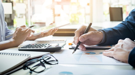 two people sit across from a table with pens and calculators in hand to plan budgets