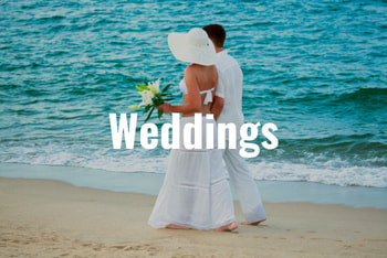 a couple wearing white walk on the beach with bright blue water behind them